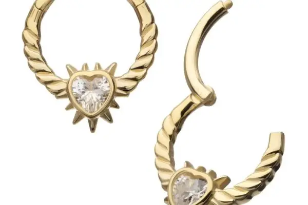 The 14K Spike Heart Clicker features gold hoops with a twisted design, a heart-shaped clear gemstone at the center, and a sunburst pattern; one earring is open, the other closed, showcasing its unique style.