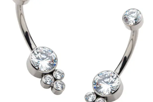 The Tri-Gem Bezel Navel Curve features a large clear central gemstone, a smaller gem at the top, and three tiny clustered gems beside the main stone in a silver-tone setting.