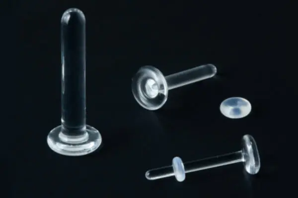 Three clear acrylic piercing retainers, including a Labret Glass Retainer, are displayed on a black surface next to a small round end piece.