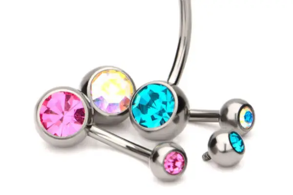 Three Double Bezel Crystal titanium belly button rings—one pink, one iridescent, and one blue—are displayed on a white background, each with curved bars and sparkling crystal gem studs. Two matching small gem ends are also shown.