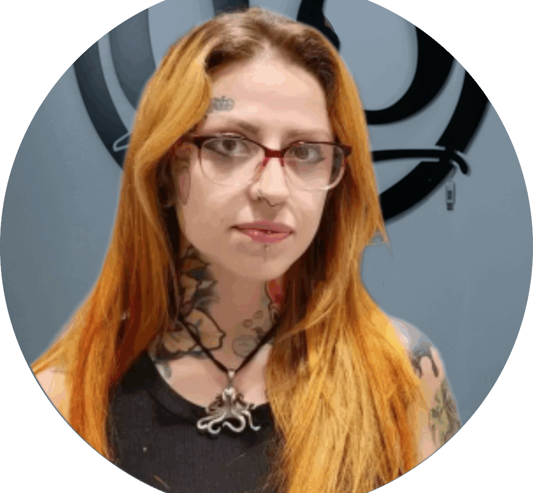 A person with long orange hair, glasses, and multiple tattoos, including on their neck and shoulders, stands in front of a gray wall with a black circular logo. They wear a black top and a necklace with an octopus pendant.