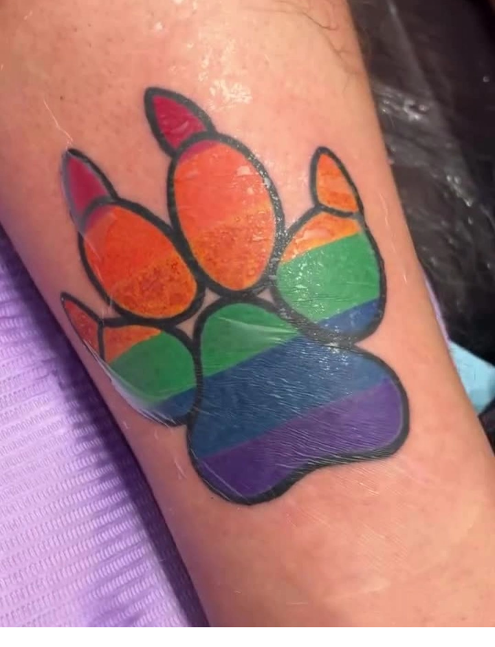 A colorful tattoo of a paw print filled with the rainbow stripes of the LGBTQ+ pride flag on a person’s forearm.
