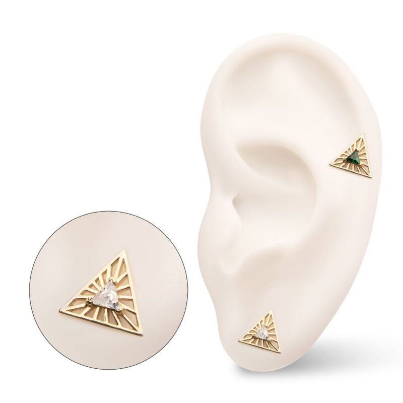 A white ear model displays two 14K Radiant Triangle with Gem earrings—one with a clear gemstone, the other green—each featuring gold geometric cutouts; an enlarged view highlights their intricate design.