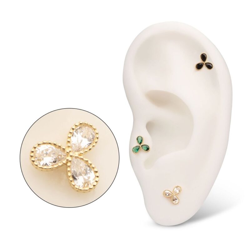 A white ear model displays three 14K 3 Petal Flower stud earrings in black, green, and clear stones with gold edges. An enlarged view shows the clear stone 14K 3 Petal Flower stud in detail.
