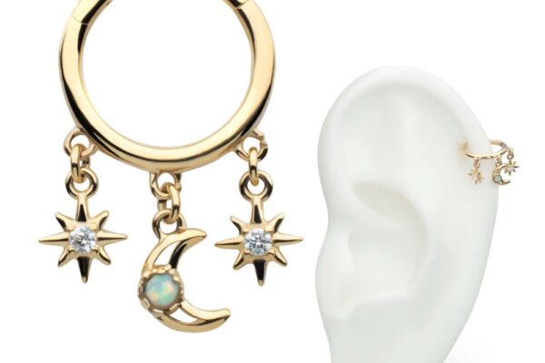 The 14K Celestial Dangle Clicker is a gold hoop earring with a crescent moon charm featuring an opal and two gemmed star charms, shown next to a model ear wearing it on the upper cartilage.