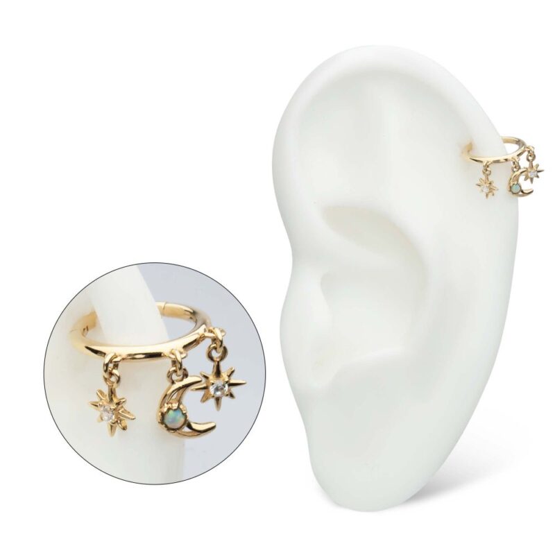 The 14K Celestial Dangle Clicker features a gold ear cuff with three charms—a star, a crescent moon with a blue gem, and a sun—displayed on a white ear model alongside a close-up of the jewelry.