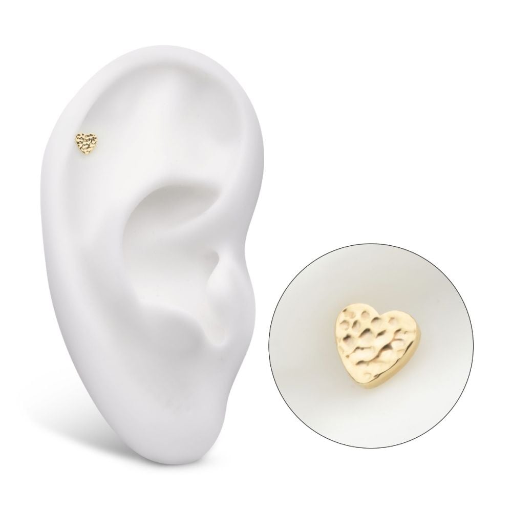 The 14K Heart – Hammered earring, a small textured gold heart-shaped stud, is displayed on a white ear model. A circular inset magnifies the intricate hammered surface of the heart design. The 14K Heart – Hammered earring, a small textured gold heart-shaped stud, is displayed on a white ear model. A circular inset magnifies the intricate hammered surface of the heart design.