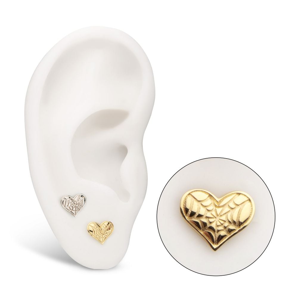 A white model ear displays two 14K Spiderweb Carved Heart earrings, one silver-toned and one gold-toned. A close-up highlights the detailed webbed design of the gold heart earring. A white model ear displays two 14K Spiderweb Carved Heart earrings, one silver-toned and one gold-toned. A close-up highlights the detailed webbed design of the gold heart earring.