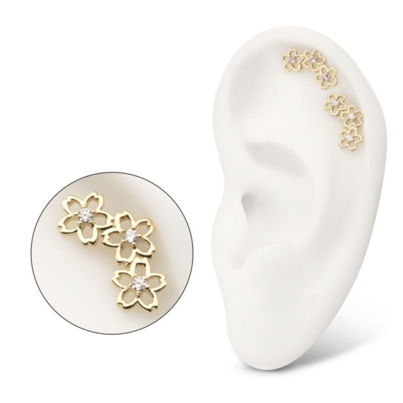 A white ear model displays three 14K Cherry Blossom Cluster earrings with gold floral shapes and central gems along the cartilage. An inset offers a close-up of these delicate earrings.