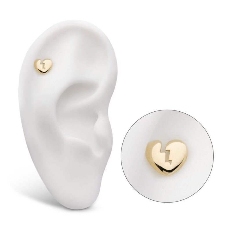 The 14K Heart Broken earring, featuring a gold heart shape with a lightning bolt crack design, is displayed on a white ear model. An inset circle highlights the earring's intricate details.