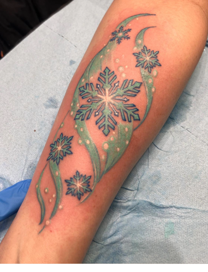 A colorful tattoo of blue snowflakes with white accents, surrounded by green swirling lines and small white dots, is inked on a persons forearm. The tattoo is fresh, and the skin around it appears slightly red.