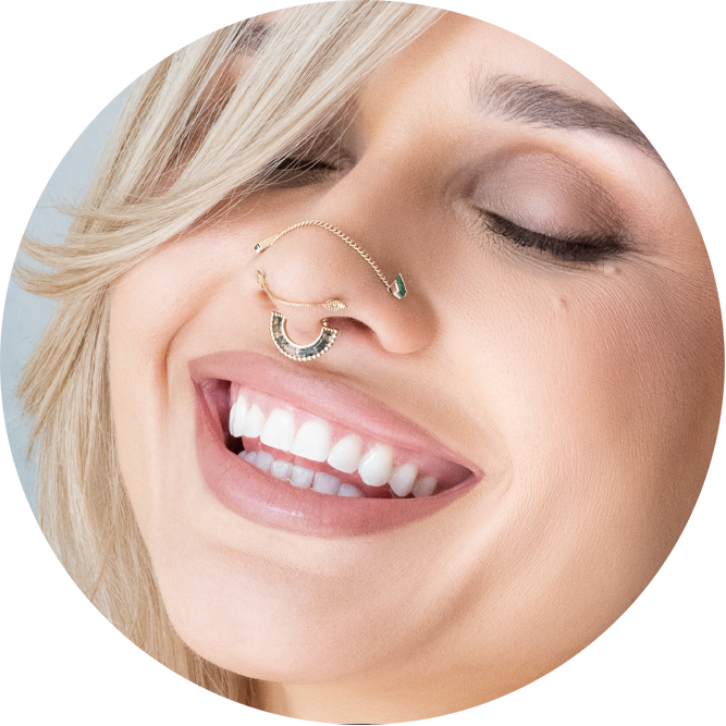 A smiling person with light skin and blonde hair wears unique nose jewelry connecting a nose ring to a septum ring, their eyes closed, teeth showing—showcasing creativity and style beyond just prices for piercing services.