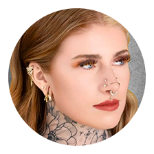 A person with long, light brown hair, multiple ear piercings, a septum and nose ring, bold makeup, and a floral neck tattoo looks to the side against a gray background—perfect for showcasing your style while reviewing our Appointment Policy.