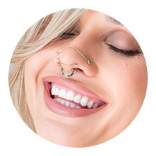 A smiling person with light hair and closed eyes shows off a septum piercing, nose ring, and bright teeth, embodying natural makeup—perfect inspiration for your next look. Be sure to check our Appointment Policy before booking!.