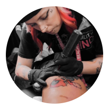 A tattoo artist with pink hair and black gloves is focused on tattooing a colorful design on a persons skin. The artist is wearing a black shirt and a lanyard.