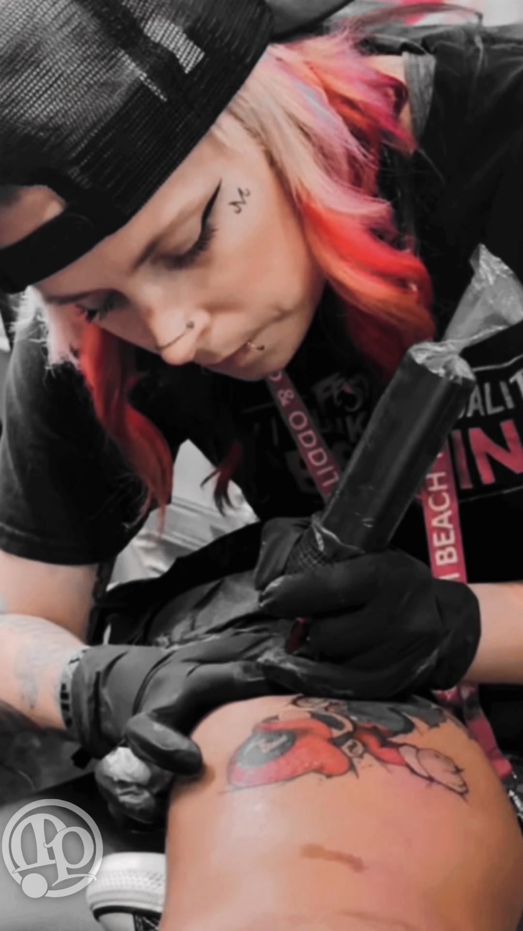 A tattoo artist with pink hair, piercings, and black gloves is focused on tattooing a colorful design. Wearing a black cap and shirt with a lanyard, she expertly applies tattoos while discussing numbing creams for client comfort.