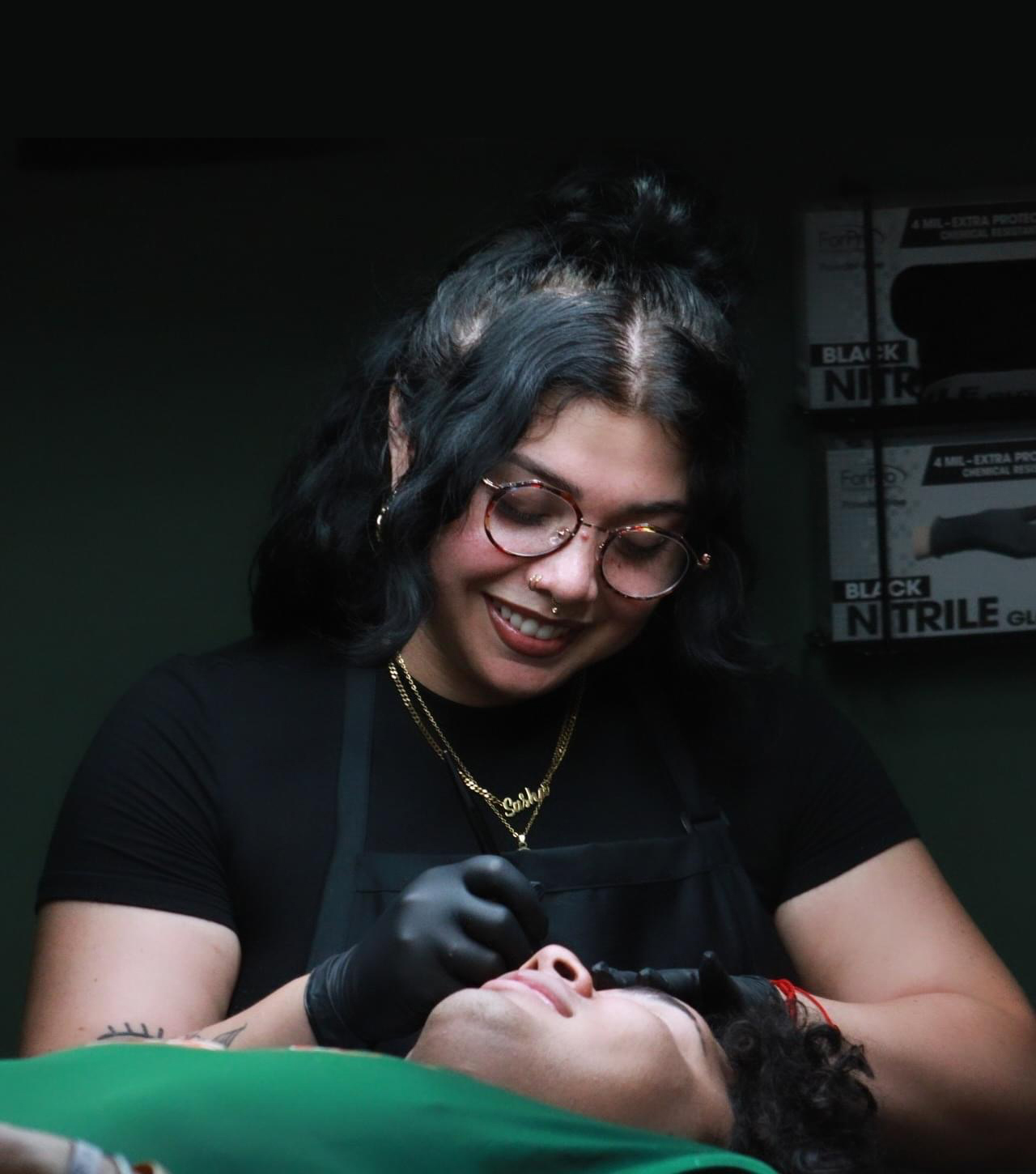 A smiling person with dark hair, glasses, and gloves is performing a beauty or cosmetic procedure on someone lying down, against a dark background with boxes labeled Nitrile Gloves.