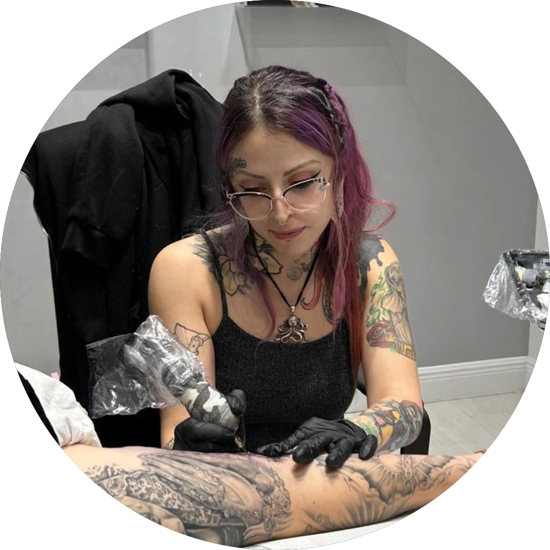 A tattoo artist with purple hair and glasses works on a client’s arm tattoo in a studio. She wears black gloves, reveals her own detailed tattoos, and keeps her equipment organized—Select Your Tattoo Artist for an inspired experience.