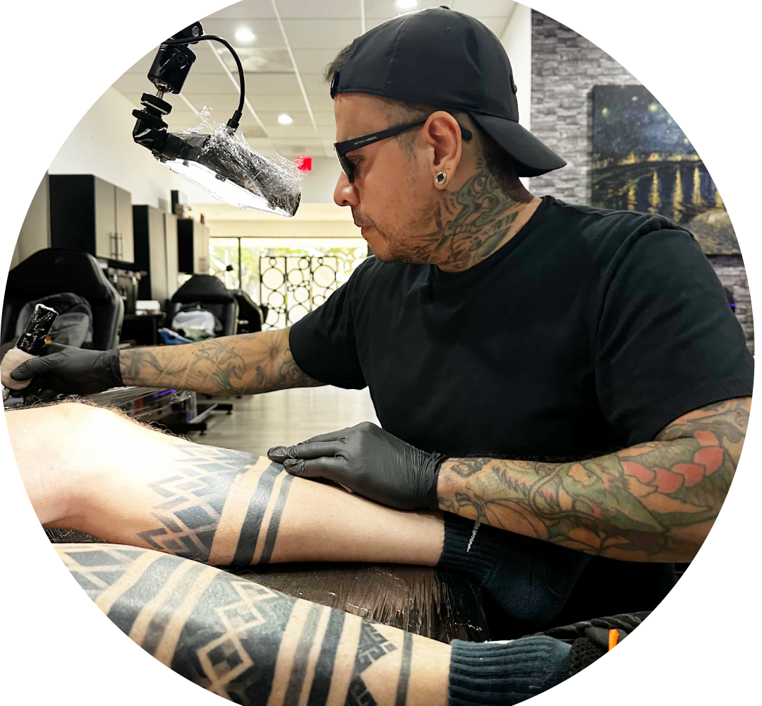 A tattoo artist with tattoos on his arms and neck works on a client’s leg tattoo in a modern studio. Select your tattoo artist as he focuses, wearing a black shirt, gloves, cap, and glasses under bright lighting.