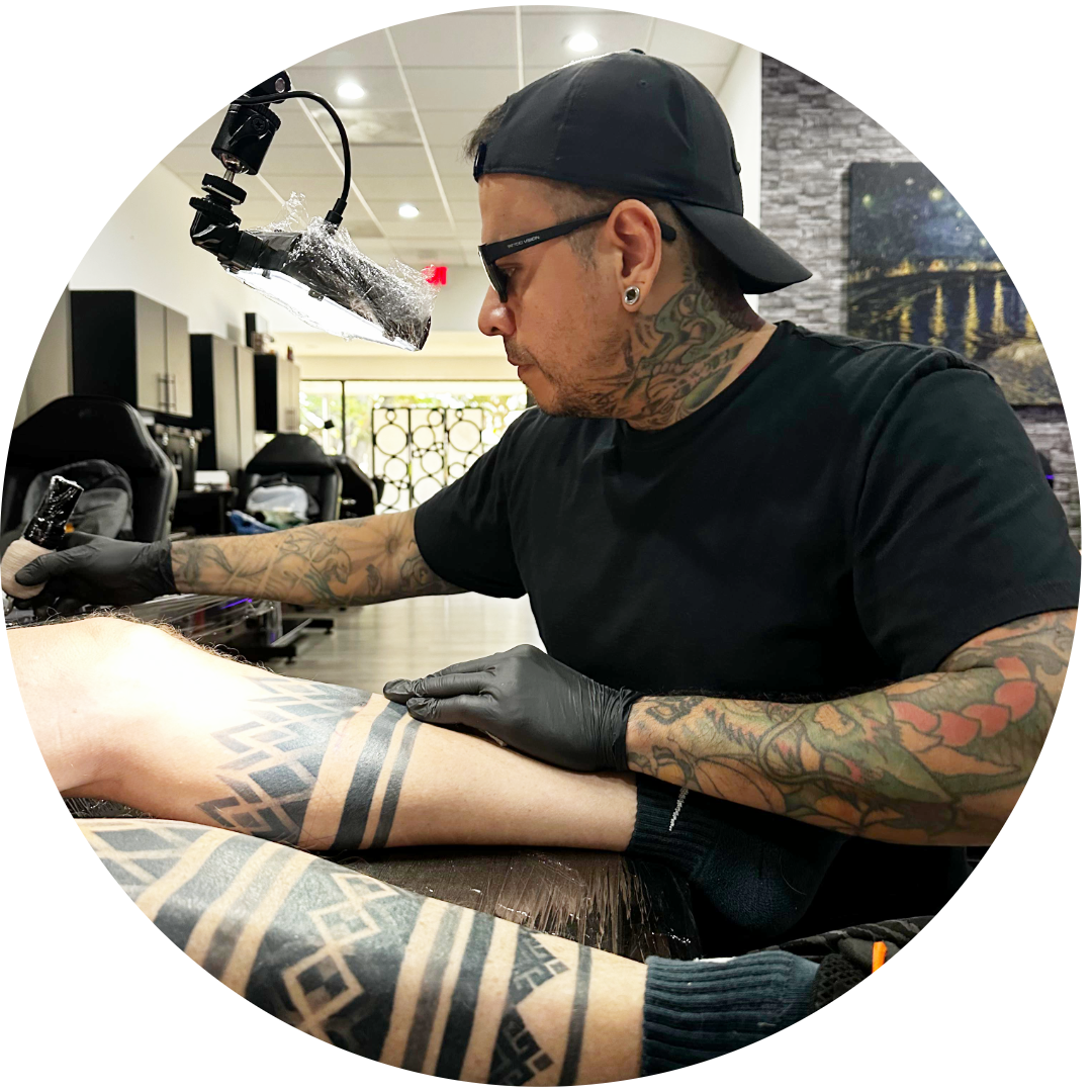 A tattoo artist with tattoos on his arms and neck works on a client’s leg tattoo in a modern studio. Select your tattoo artist as he focuses, wearing a black shirt, gloves, cap, and glasses under bright lighting.