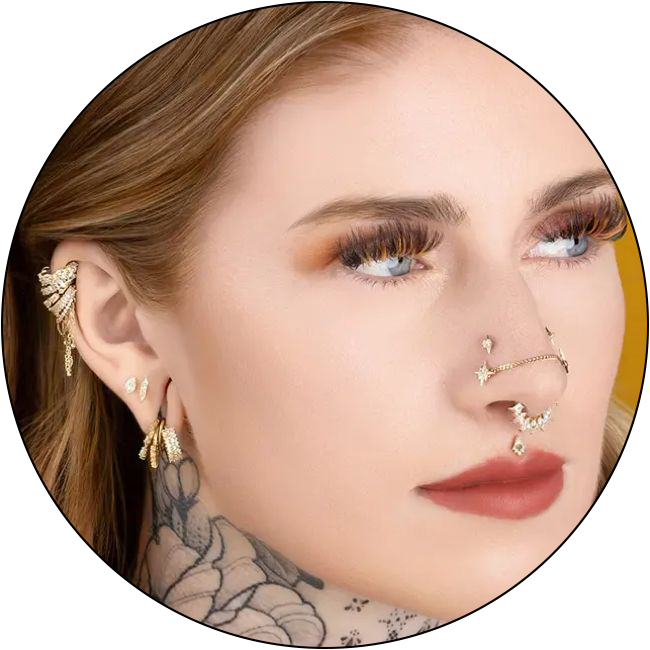 A woman with blue eyes and long lashes wears multiple gold earrings, a nose ring, and a nose chain. She has a floral neck tattoo and glances to the side against a yellow background, embodying elegance with an eye for detail—just like our Appointment Policy.