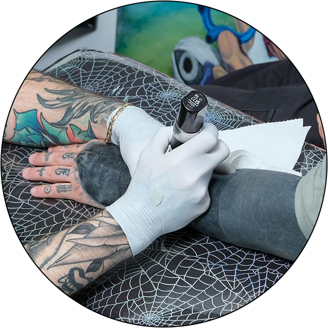 A tattoo artist with colorful arm tattoos uses a tattoo machine to ink a client’s mostly black forearm. The artist, following the studio’s Appointment Policy, wears white gloves and rests the arm on a web-patterned surface.