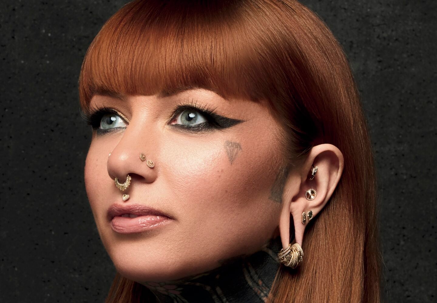 Meet Your Piercer: A woman with straight red hair and bangs, multiple facial and ear piercings, bold black eyeliner, a small heart tattoo on her cheek, and a tattooed neck stands against a dark textured background.