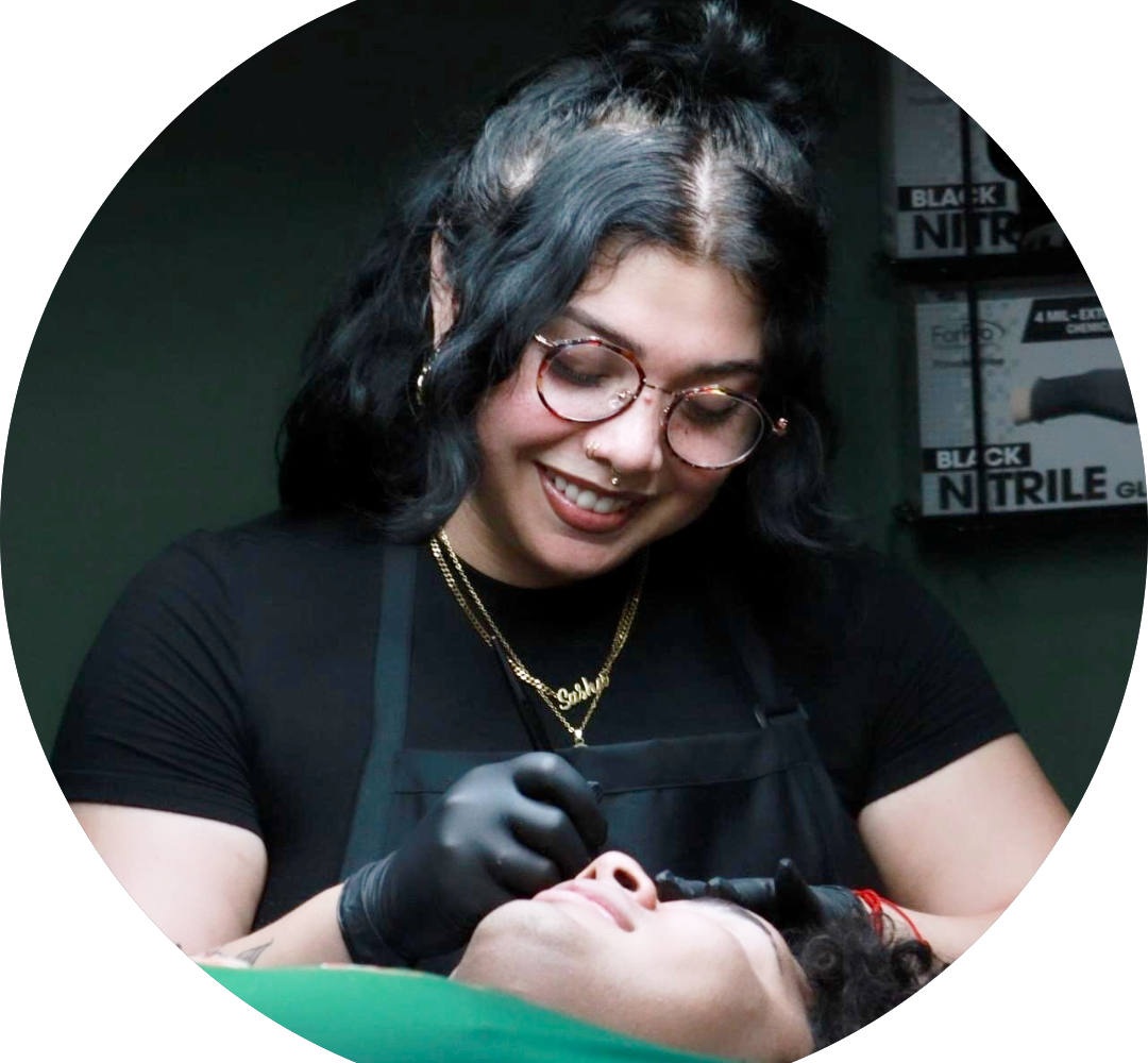 A person with glasses and black gloves smiles while performing a cosmetic procedure on another person lying down, in a room with boxes labeled Nitrile Gloves in the background.