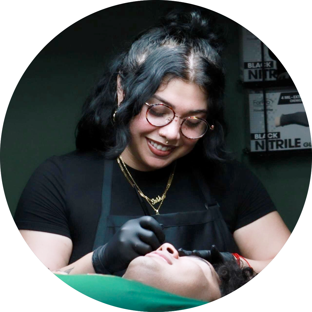A person with glasses and black gloves smiles while performing a cosmetic procedure on another person lying down, in a room with boxes labeled Nitrile Gloves in the background.