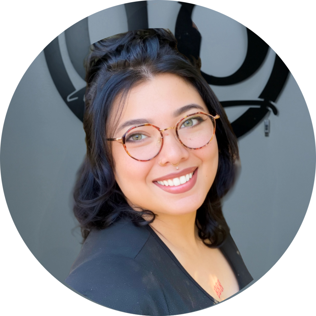 A person with medium-length dark hair, round glasses, and a nose ring smiles at the camera. They are wearing a black top and standing in front of a gray wall with a circular black design.