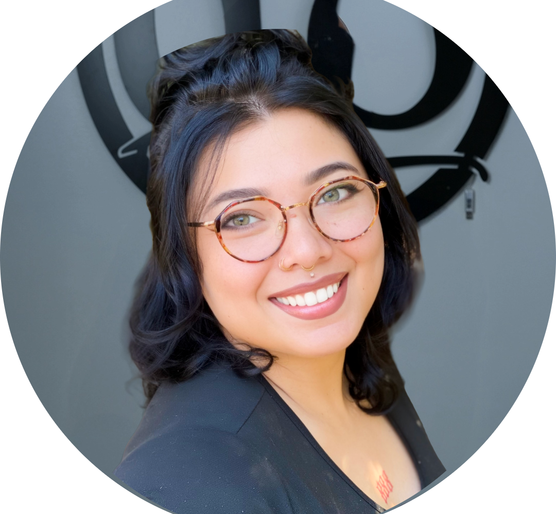 A person with medium-length dark hair, round glasses, and a nose ring smiles at the camera. They are wearing a black top and standing in front of a gray wall with a circular black design.