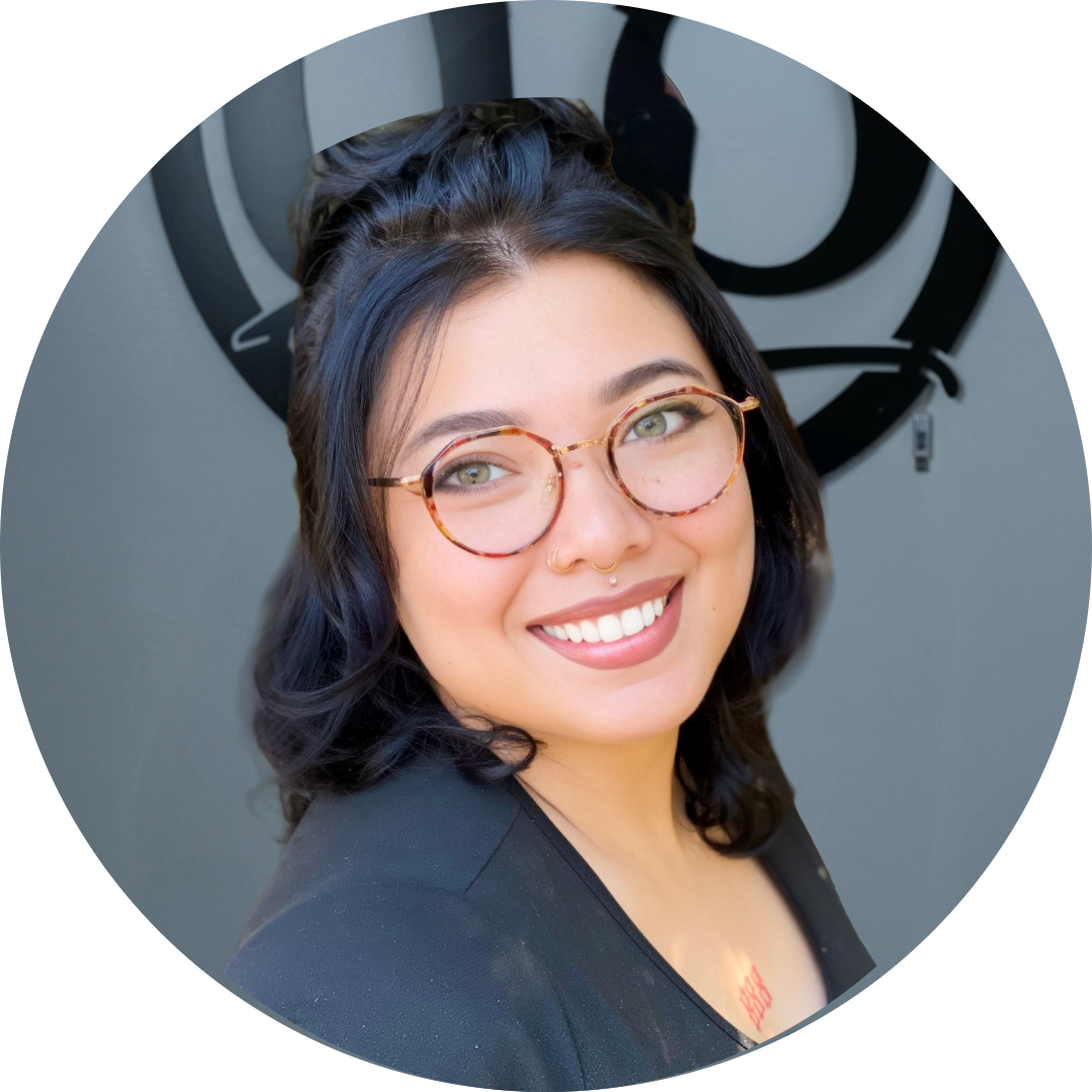 A person with medium-length dark hair, round glasses, and a nose ring smiles at the camera. They are wearing a black top and standing in front of a gray wall with a circular black design.
