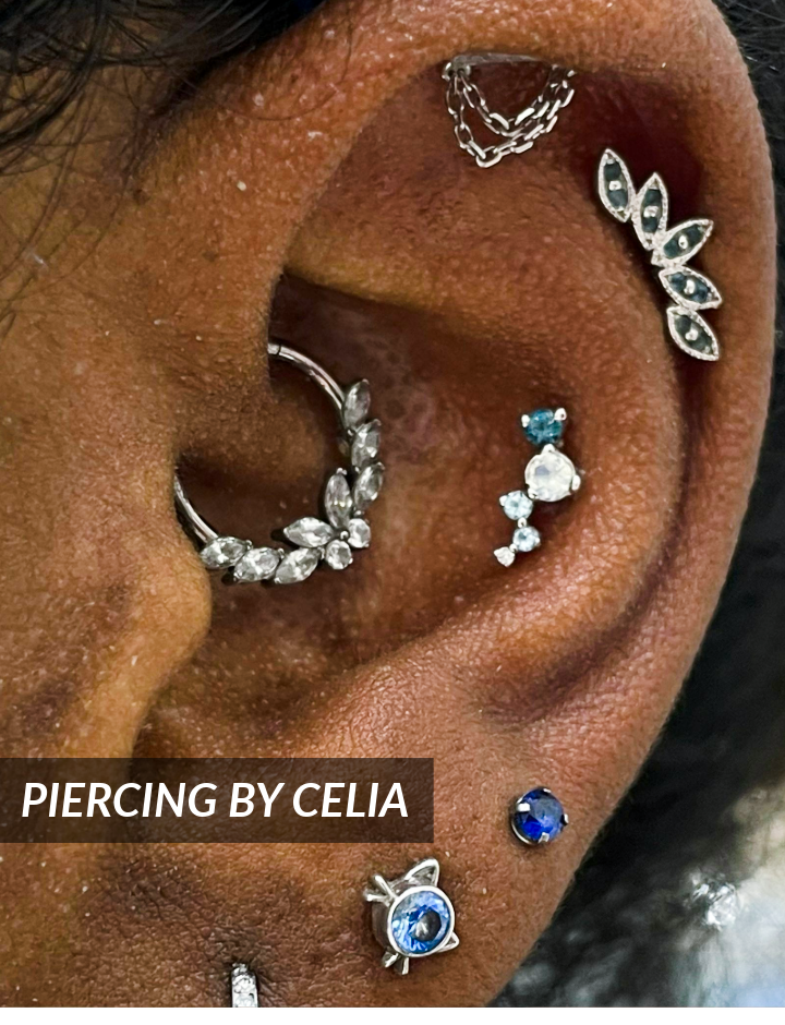 A close-up of an ear with multiple piercings featuring decorative silver and blue gemstone jewelry. Text on the image reads PIERCING BY CELIA.