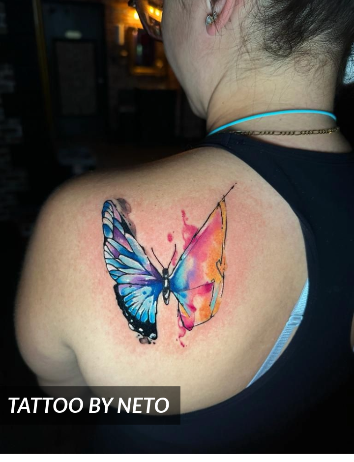 A colorful butterfly tattoo with blue and pink watercolor effects is inked on a woman’s upper back. She is wearing a black tank top, with the tattoo artist’s name Tattoo by Neto in the corner.