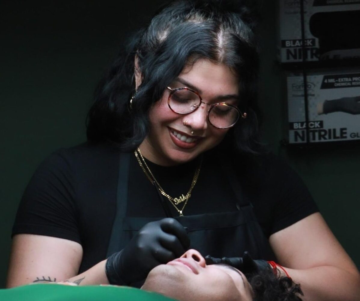 Meet Skie, a talented tattoo artist with glasses and dark hair, wearing black gloves and an apron. She smiles while performing a cosmetic procedure on a person lying down, with boxes of black nitrile gloves visible in the background.