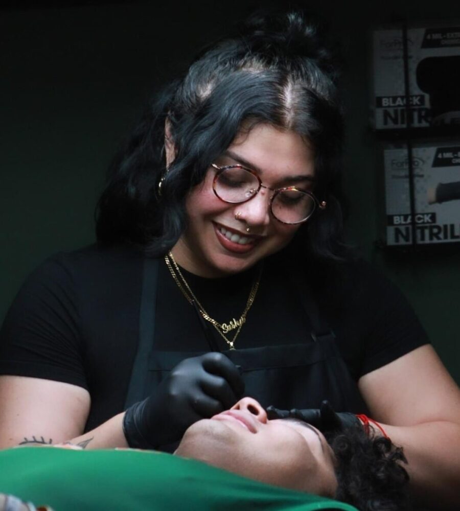 Meet Skie, a talented tattoo artist with glasses and dark hair, wearing black gloves and an apron. She smiles while performing a cosmetic procedure on a person lying down, with boxes of black nitrile gloves visible in the background.