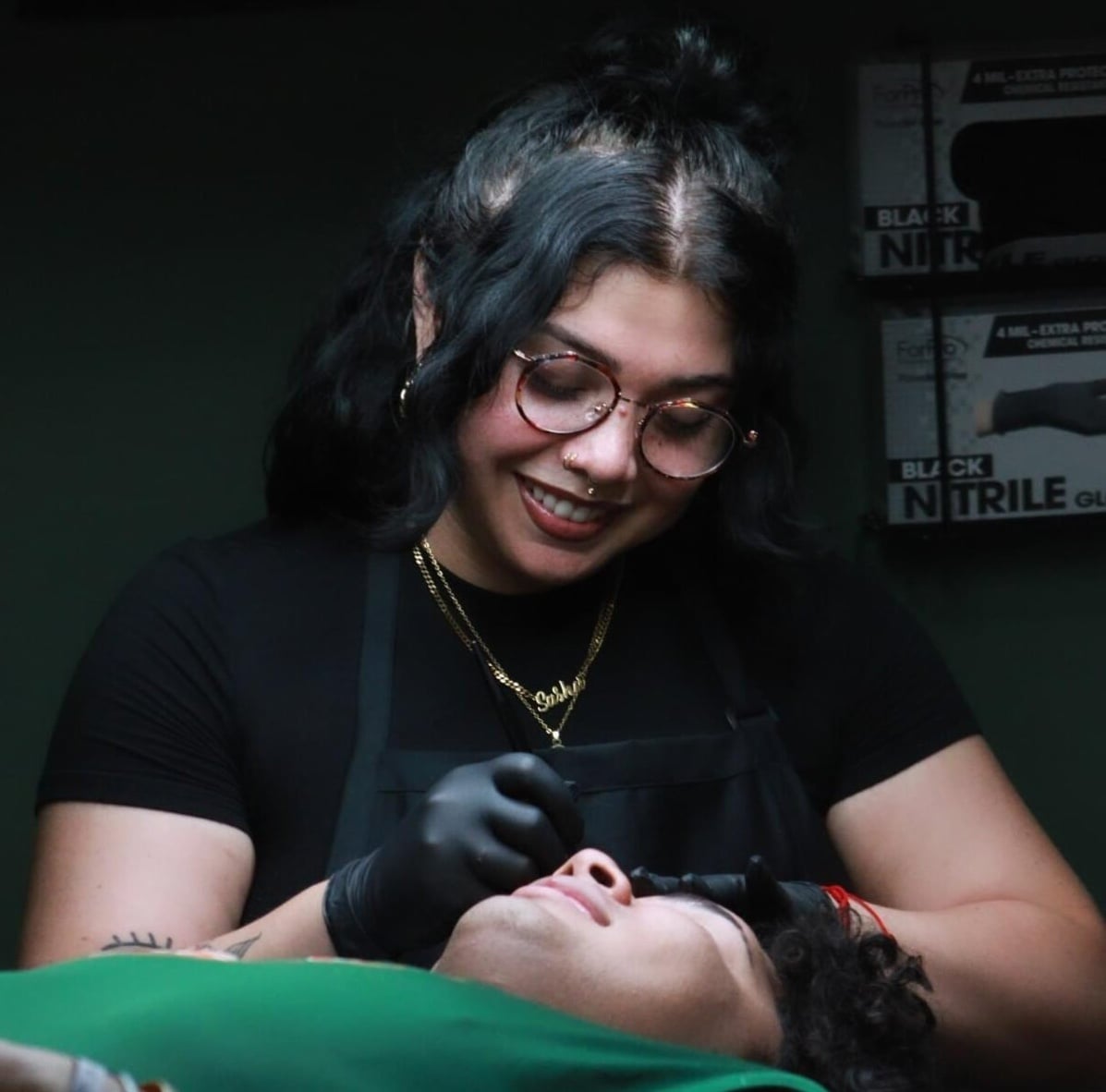 Meet Skie, a talented tattoo artist with glasses and dark hair, wearing black gloves and an apron. She smiles while performing a cosmetic procedure on a person lying down, with boxes of black nitrile gloves visible in the background.