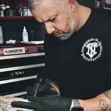 Meet Andrea, a tattoo artist with gray hair and a black t-shirt, who focuses intently while tattooing a client's arm, wearing black gloves and using a tattoo machine in the studio.