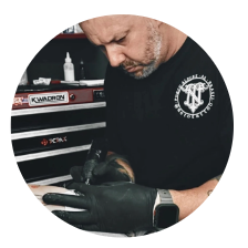 A tattoo artist wearing black gloves and a black T-shirt works intently on a tattoo, holding a tattoo machine over a client’s skin—reminding you to Select Your Tattoo Artist carefully—with a tool chest of supplies in the background.