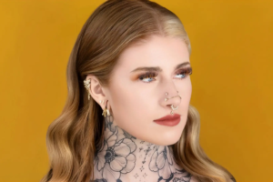 A woman with long wavy hair and blue eyes stares to the side. She has floral tattoos from tattooing, multiple gold body jewelry earrings, and several nose piercings. The background is a solid yellow color.