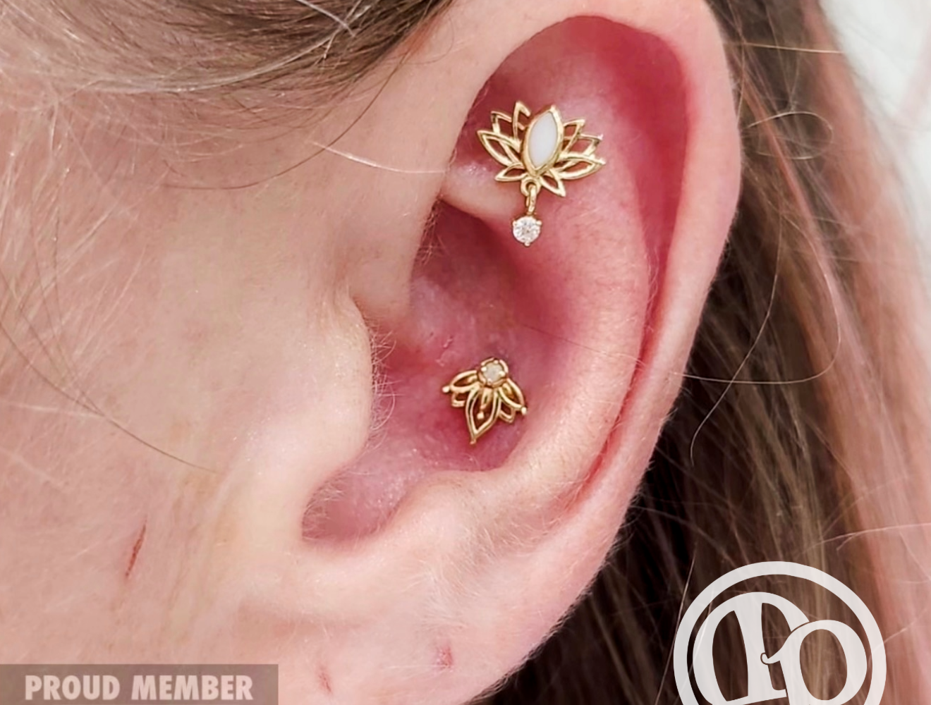 A close-up of an ear with two gold lotus flower-shaped piercings—one at the daith and one at the conch. APP membership and logo marks appear in the corners, ideal for those interested in both tattoos and quality numbing creams.