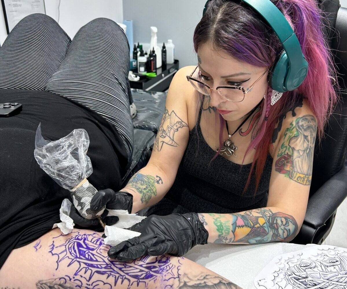 Meet Andrea, a tattoo artist with purple hair and green headphones, carefully inking a client’s thigh. She wears gloves and focuses on a purple stencil design, with tattoo equipment and bottles arranged on the table nearby.