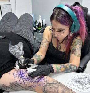 Meet Andrea, a tattoo artist with purple hair and green headphones, carefully inking a client’s thigh. She wears gloves and focuses on a purple stencil design, with tattoo equipment and bottles arranged on the table nearby.
