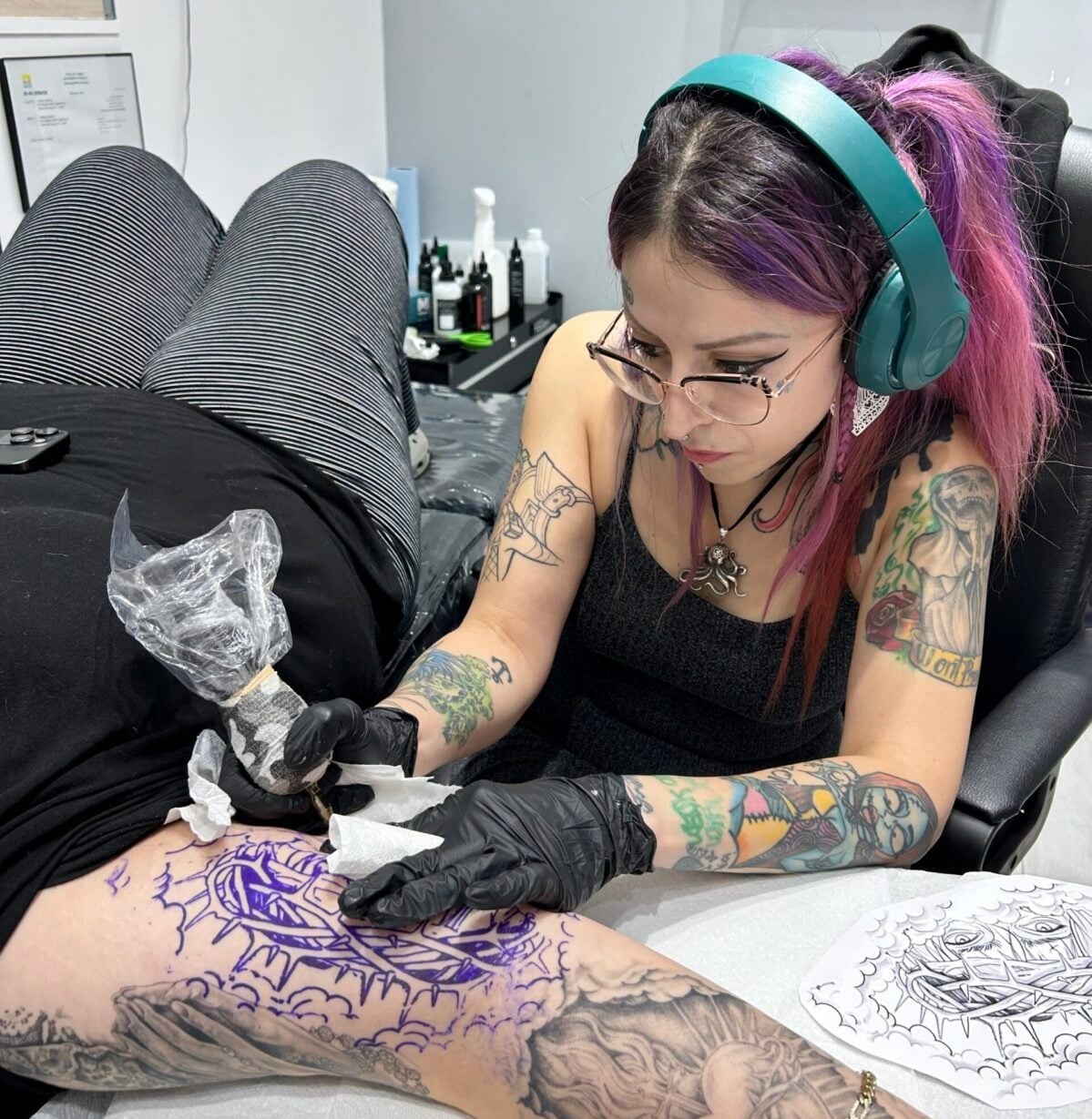 Meet Andrea, a tattoo artist with purple hair and green headphones, carefully inking a client’s thigh. She wears gloves and focuses on a purple stencil design, with tattoo equipment and bottles arranged on the table nearby.