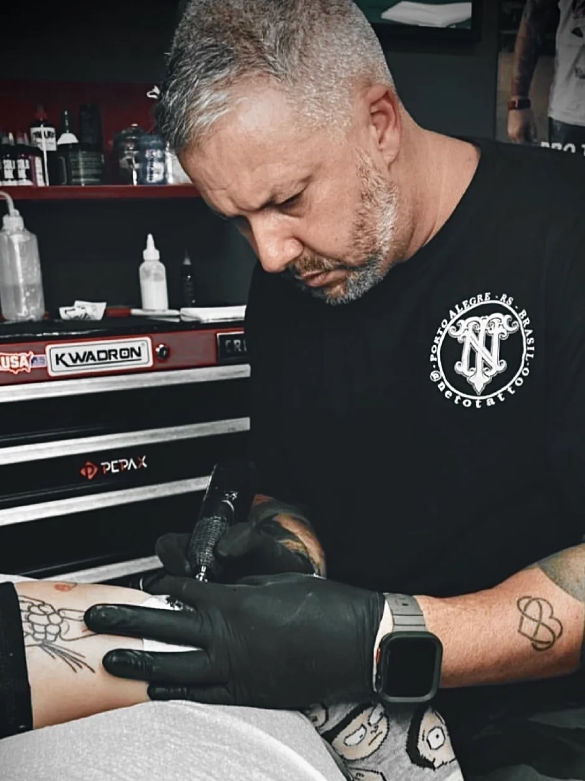 A tattoo artist with short gray hair and a beard is focused on tattooing a persons forearm. He wears black gloves and a black shirt, working in a studio with tattoo equipment and supplies visible in the background.