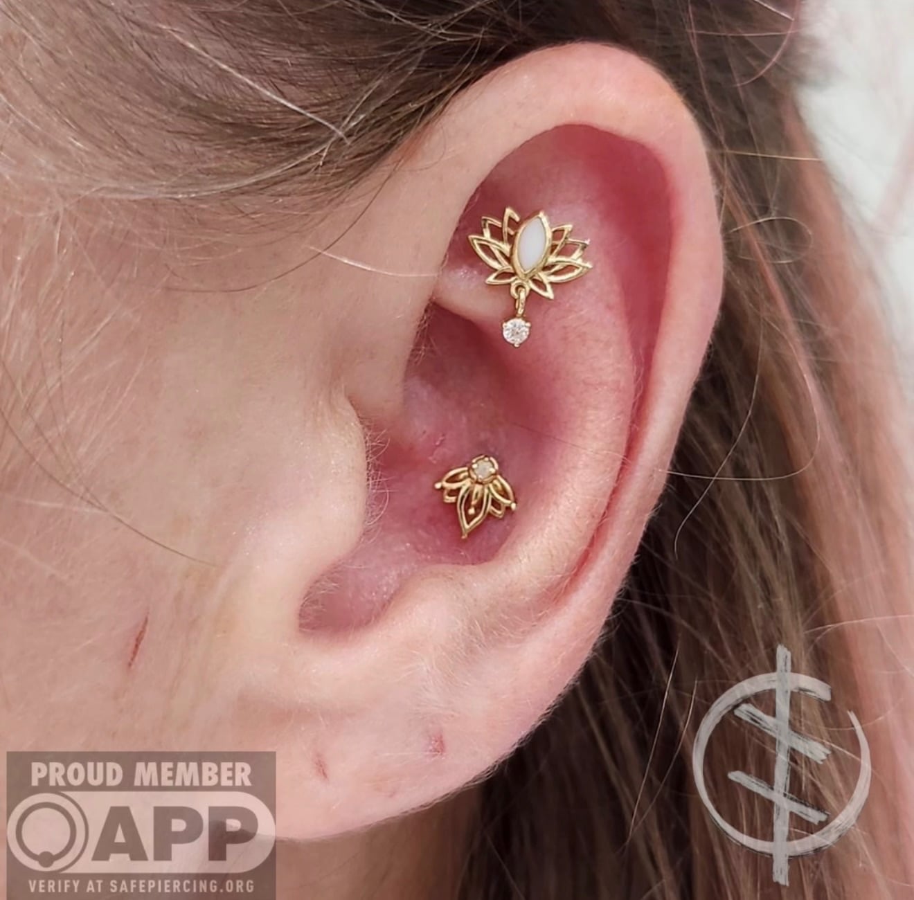 A close-up of a person's ear with two gold lotus flower piercings, one in the upper cartilage and one in the conch—perfect inspiration for tattoo and piercing lovers. The image features a Proud Member APP badge and a circular logo in the corners.