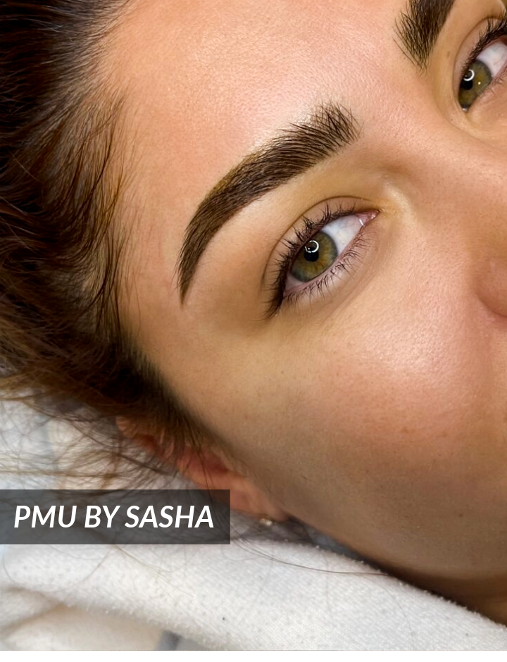 Close-up of a womans face showing well-groomed, defined eyebrows with a natural look. Text overlay reads PMU BY SASHA.