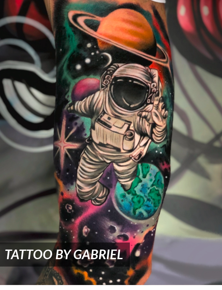 Colorful tattoo of an astronaut floating in space with planets, stars, and a swirling galaxy in the background. The tattoo is on a forearm. Text at the bottom reads TATTOO BY GABRIEL.