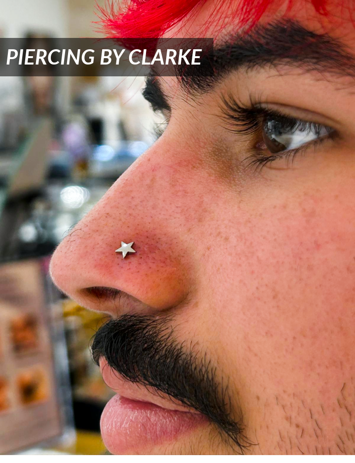 A person with red hair and a mustache has a star-shaped nose stud piercing. The text PIERCING BY CLARKE appears at the top left of the image. The background is blurred.