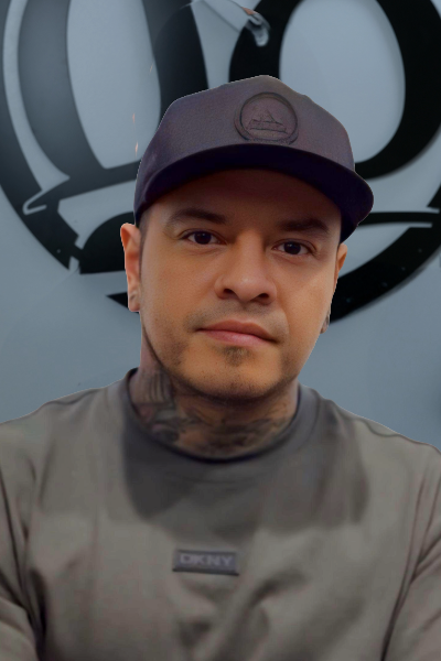 A man with a neutral expression wears a dark cap and a grey DKNY shirt, with tattoos visible on his neck. The background features large, stylized black and white lettering.